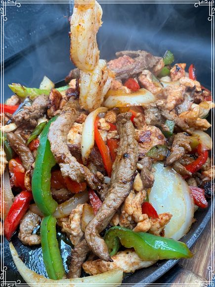 Sizzling fajitas with steak, chicken, shrimp, peppers, and onions on a hot skillet, with steam rising.