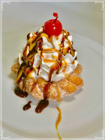 Fried dough pastry with whipped cream, caramel, chocolate drizzle, and a cherry on white plate.