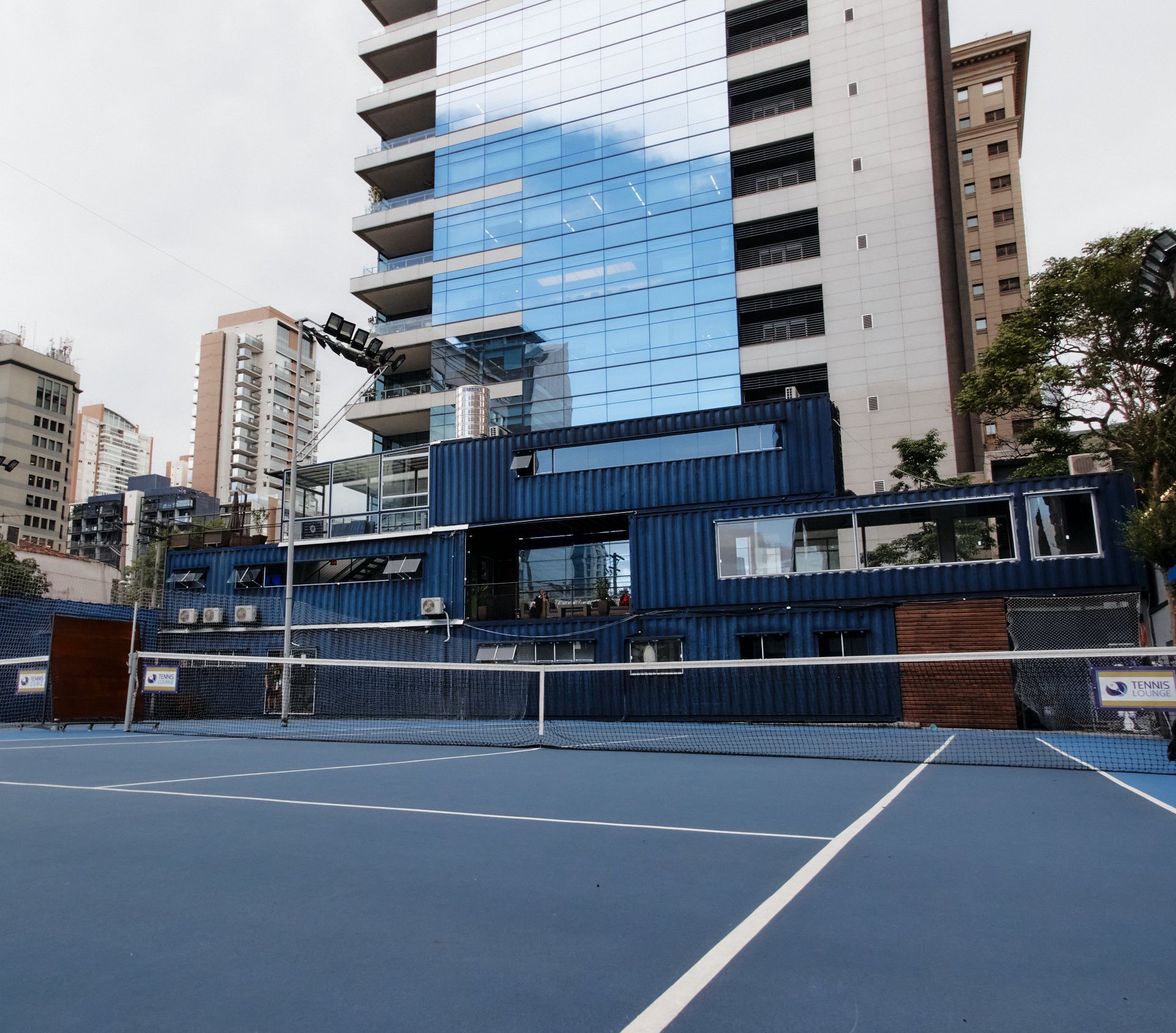 Quadra de tênis azul em frente a um edifício feito de contêineres azuis, com uma moderna torre de vidro ao fundo.