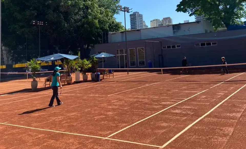 Uma quadra de tênis com dois jogadores, um de azul caminhando em direção à rede, o outro do outro lado, com prédios no fundo.