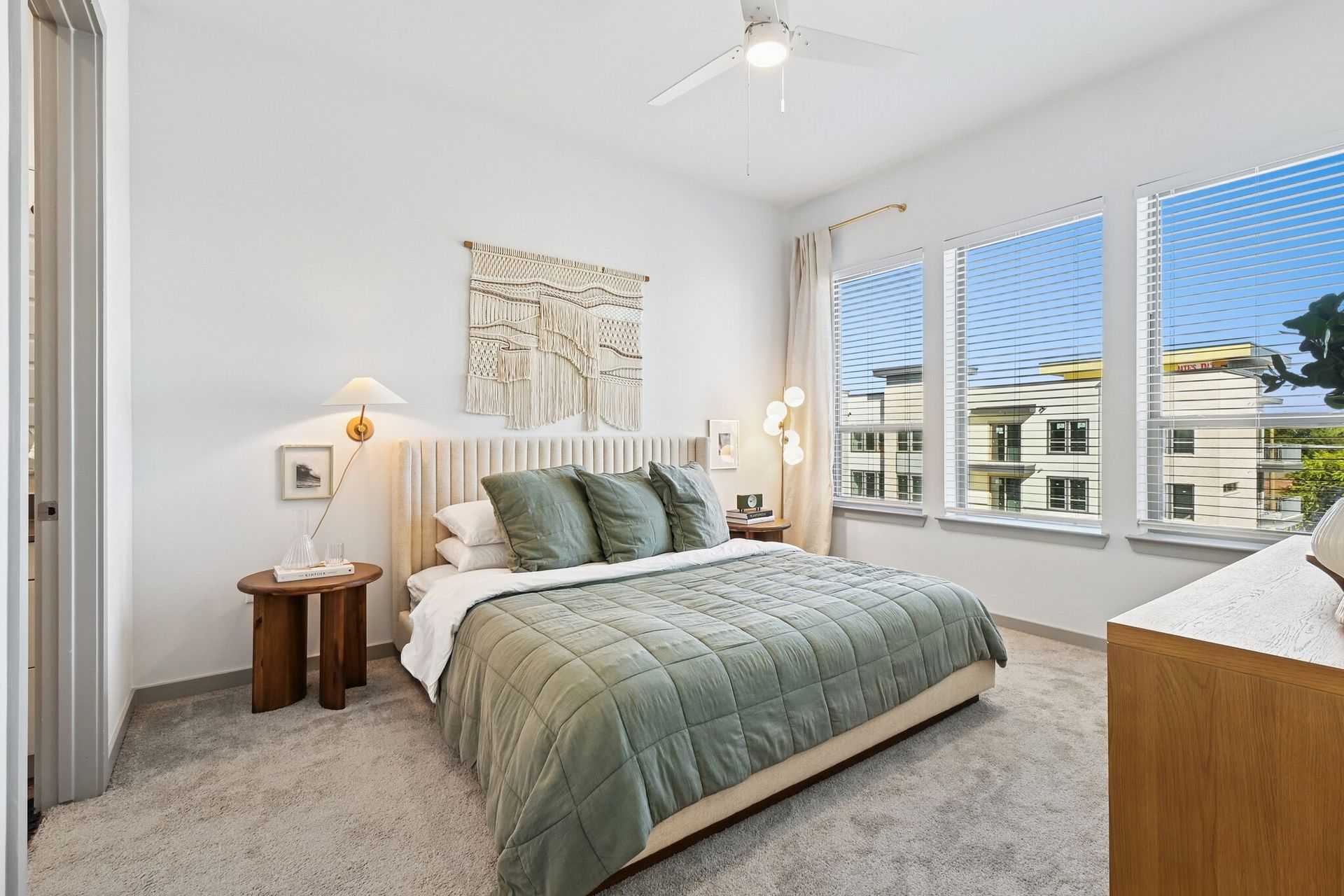 Bright bedroom in a modern apartment with a plush bed, sage bedding, and large windows.