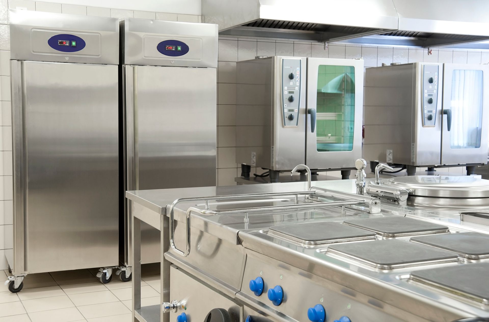 Commercial kitchen with stainless steel appliances: refrigerator, ovens, stovetop, and exhaust hood.