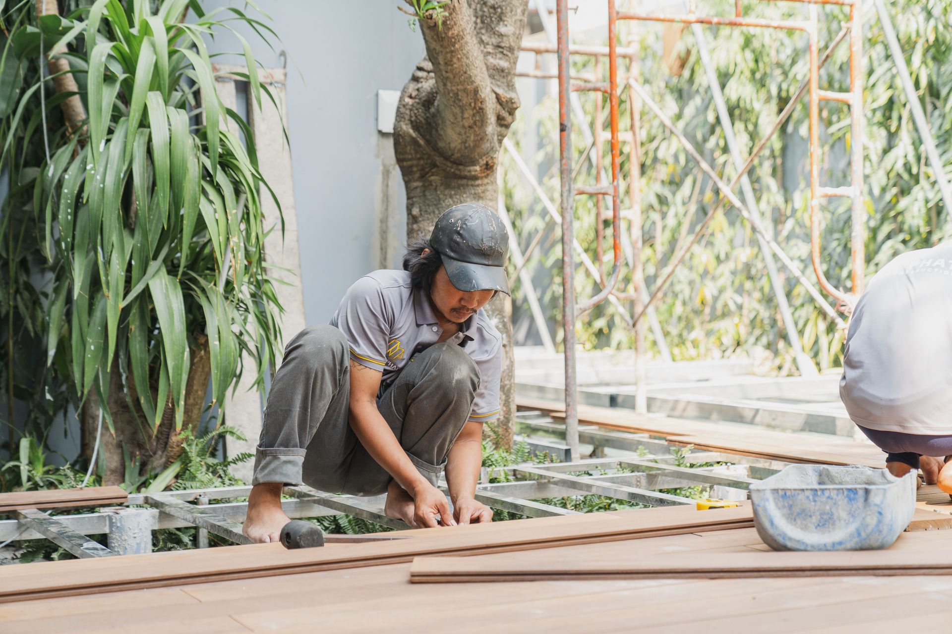 Worker crouching on a wooden deck under construction, measuring boards with plants and scaffolding nearby