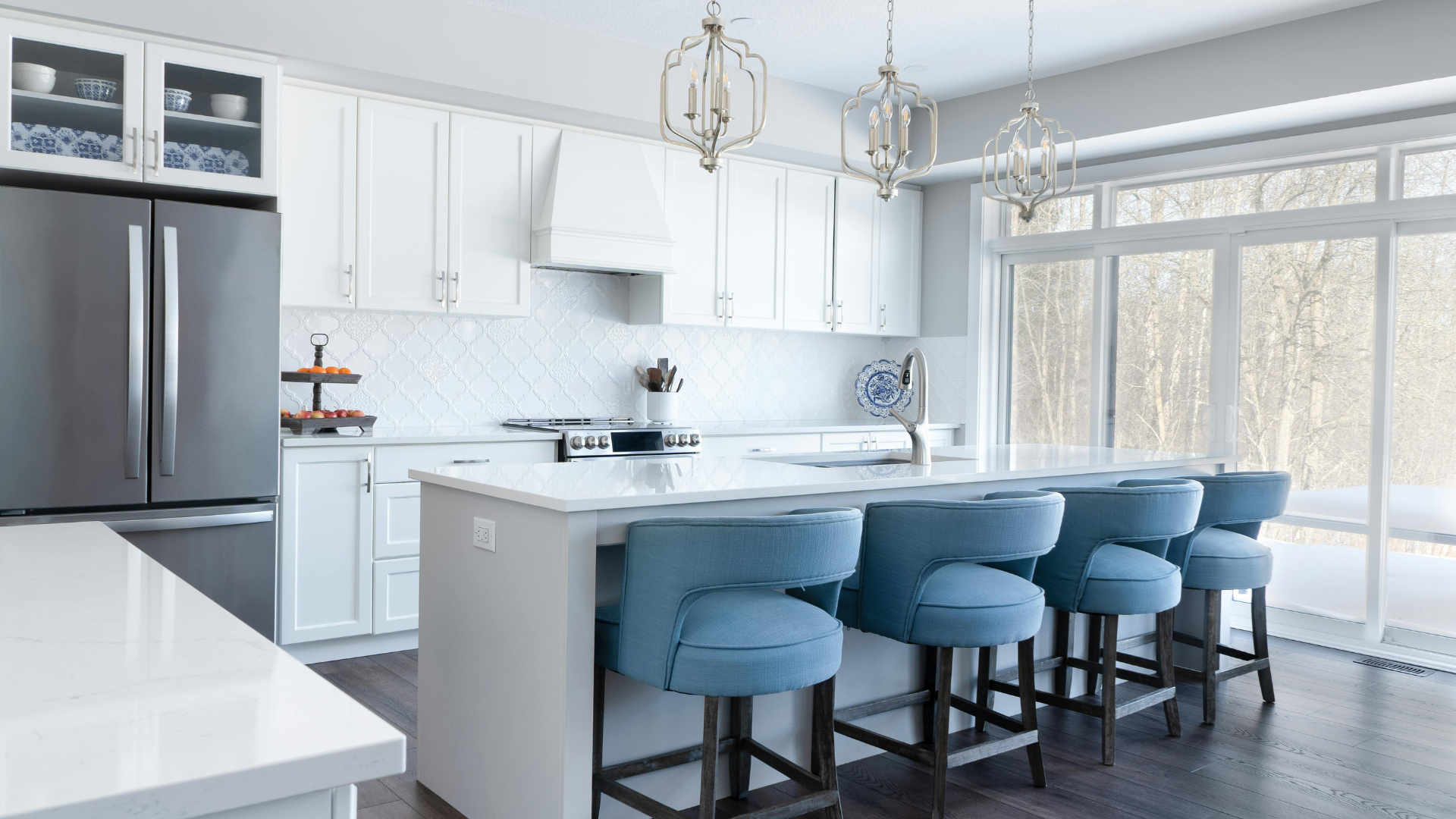 Bright modern kitchen with white cabinets, marble island, stainless fridge, and blue barstools by large windows