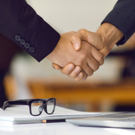 Two people shaking hands over a desk with eyeglasses and a notebook