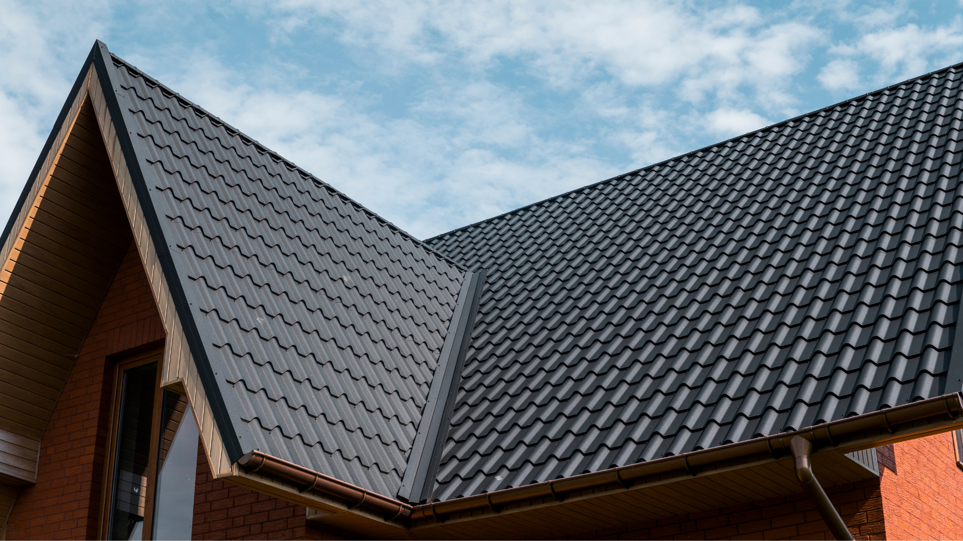 Dark gray tiled roof on a brick house under a partly cloudy sky