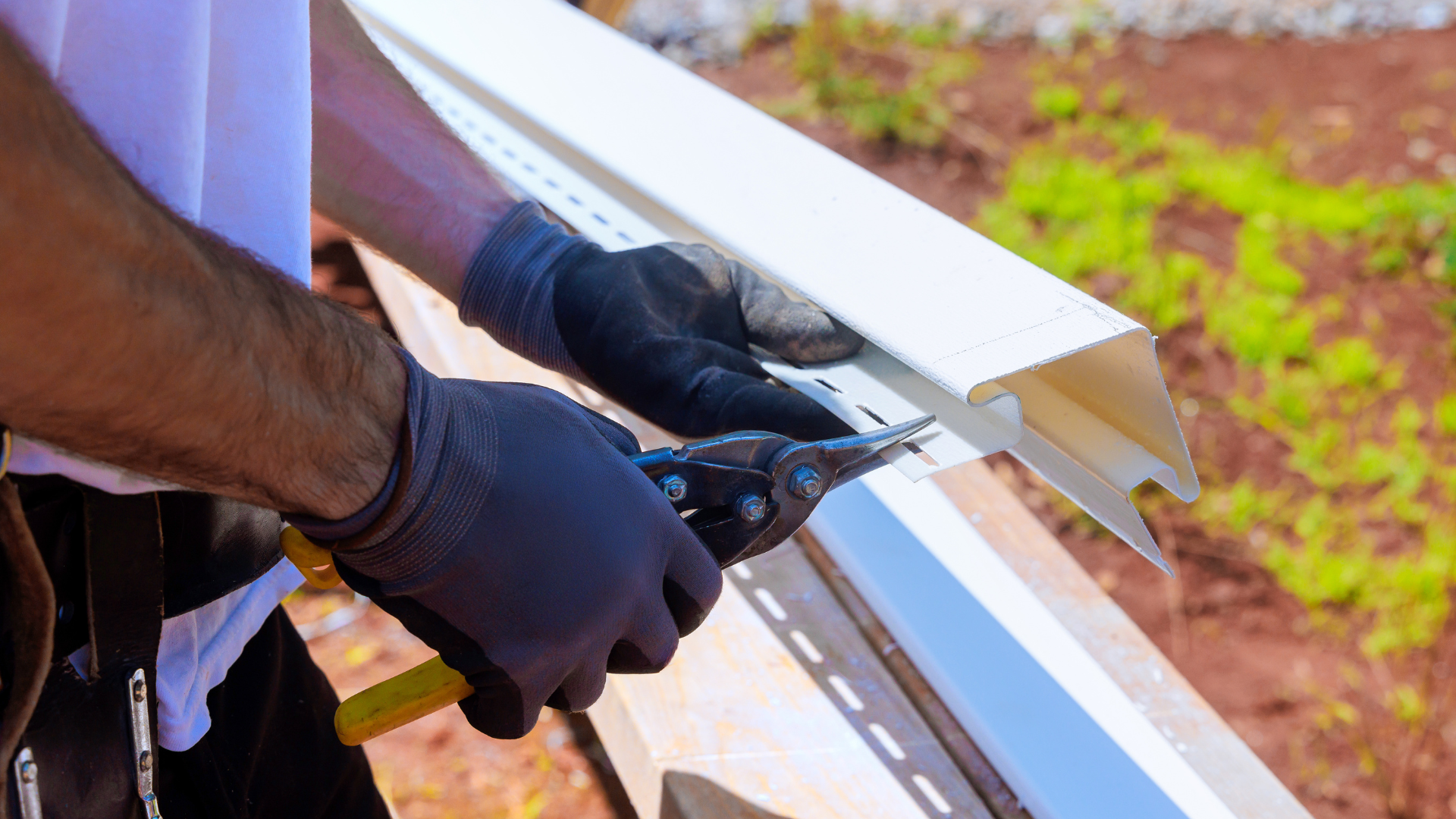 Gloved worker cutting white metal siding with snips outdoors on a roof edge