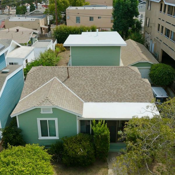 Aerial view of a green house with a beige roof in a dense residential neighborhood.