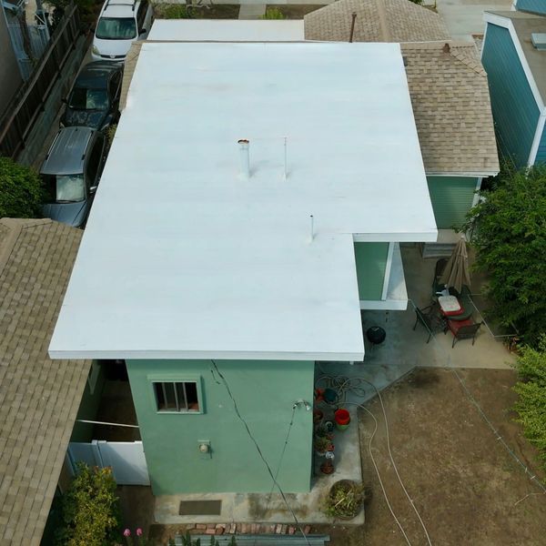 Aerial view of a house with a large white roof, mint-green walls, and surrounding yard.