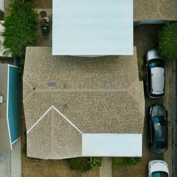 Aerial view of a tan-roofed house with parked cars along the driveway and a pool beside the home