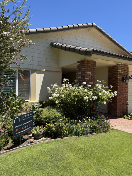 Single-story beige house with brick porch, white flowers, and a small garden sign in front yard