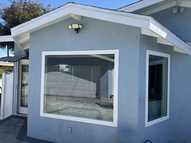 Small gray stucco building with large windows and a white roof trim in daylight