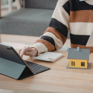Person using a laptop at a wooden desk with a notebook and a small house model