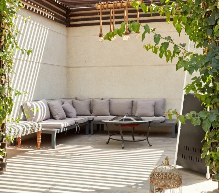 Covered patio with gray sectional seating, striped rug, and leafy vines framing the space