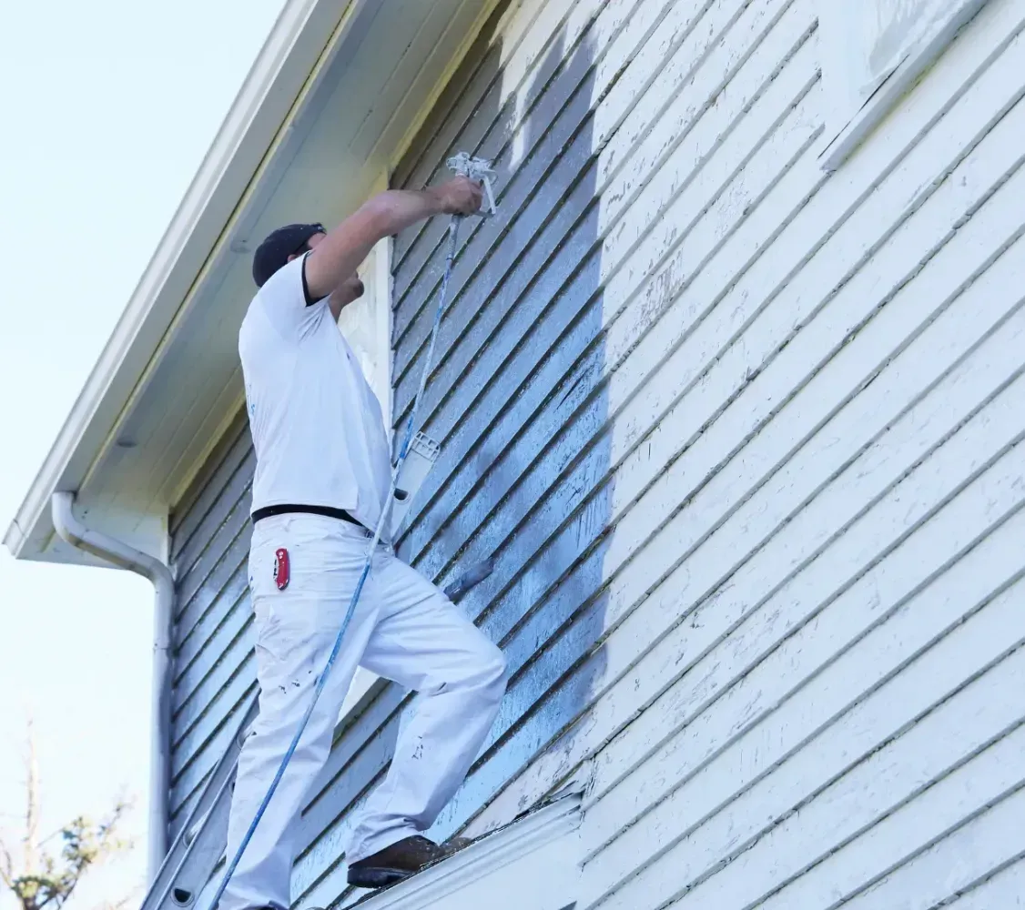 Person painting the exterior of a white house while standing on a ladder.