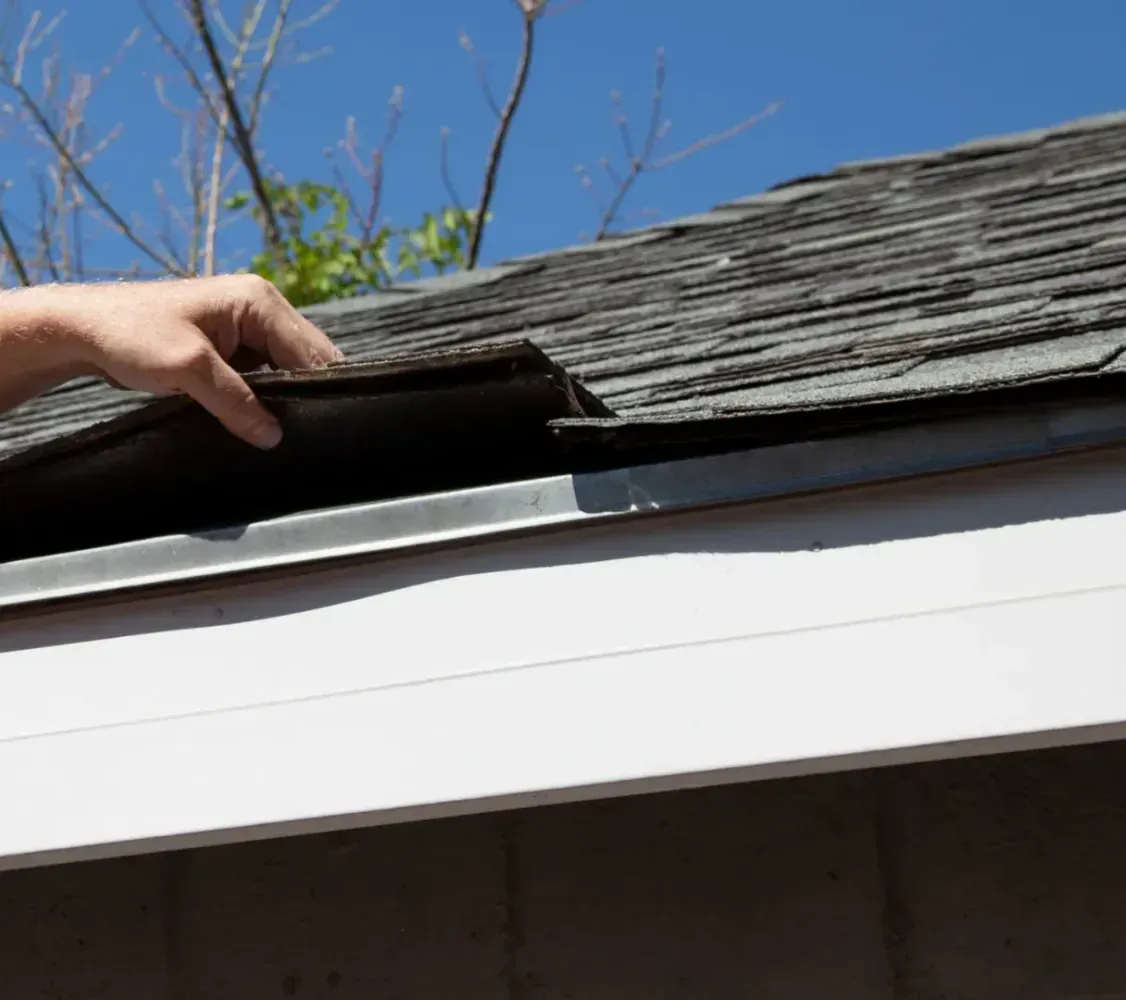 Hands lifting a shingle on a roof edge to inspect the flashing beneath