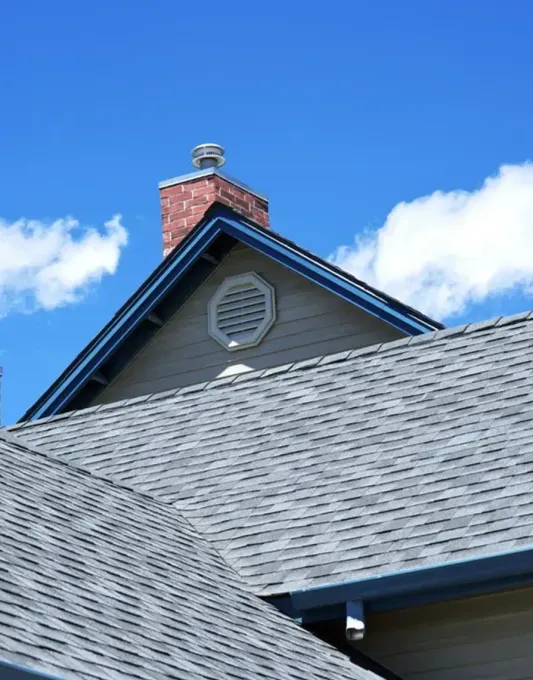 Blue-gray shingled house roof with a brick chimney under a bright blue sky and clouds