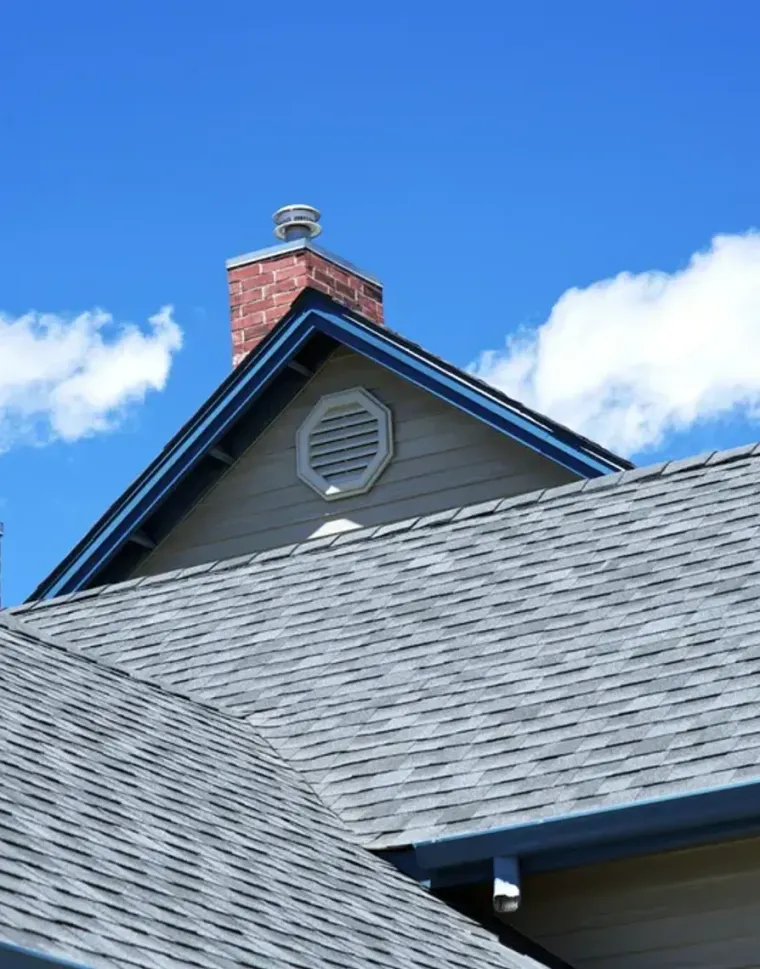Blue-gray shingled house roof with a brick chimney under a bright blue sky and clouds