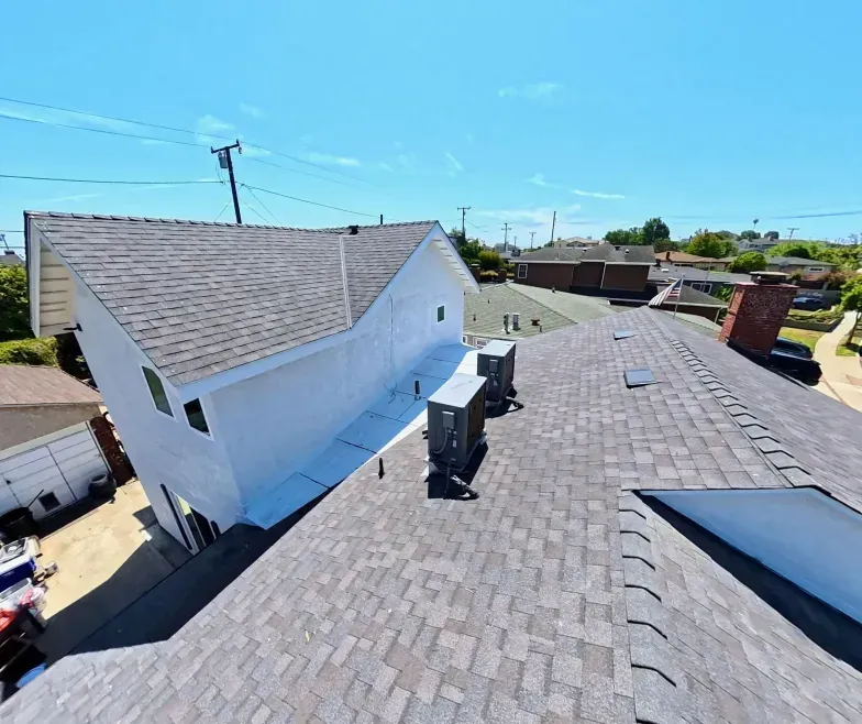 Rooftop with gray shingles and vents beside a white building under a clear blue sky.