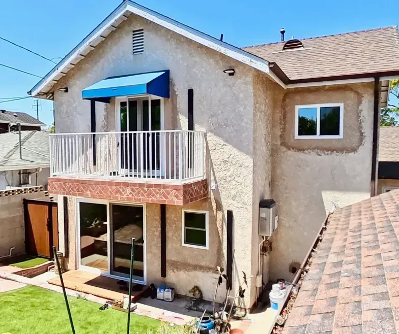 Two-story stucco house with a balcony, blue awning, and fenced backyard under a clear sky