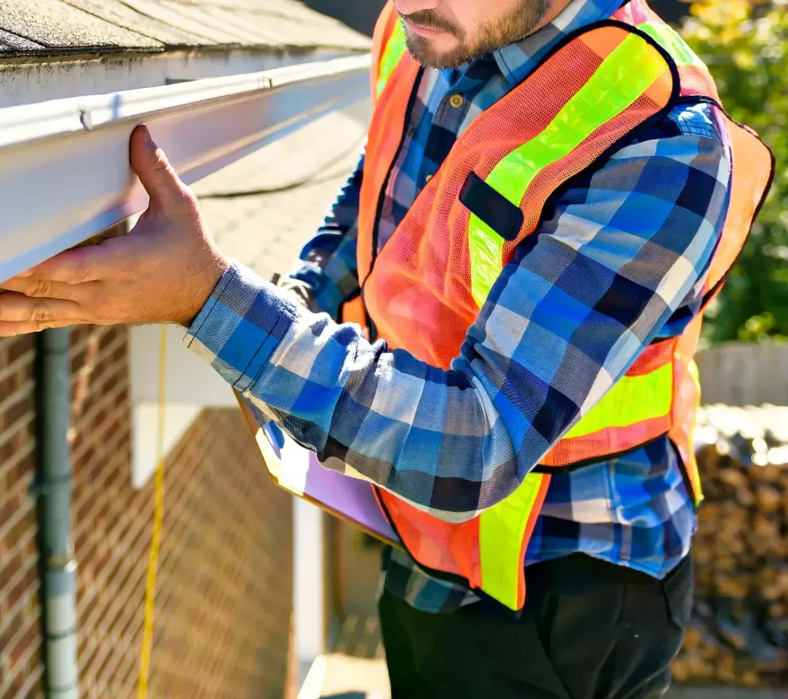 Worker in orange safety vest inspecting roof gutters on a brick house