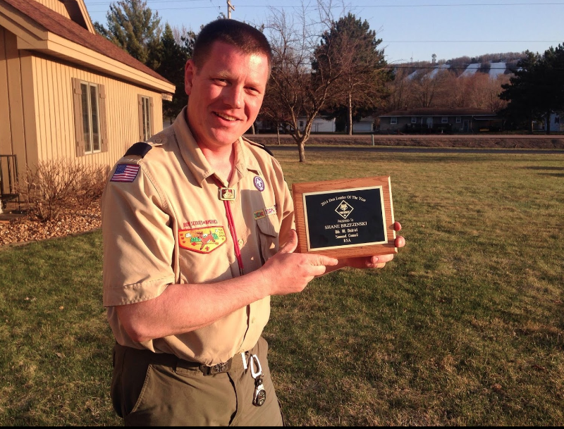 A scout in uniform stands outdoors smiling while holding a wooden plaque.