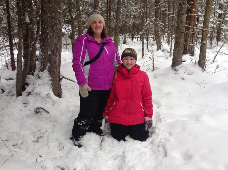 Two people in winter jackets and beanies smile while standing and kneeling in a snow-covered forest.