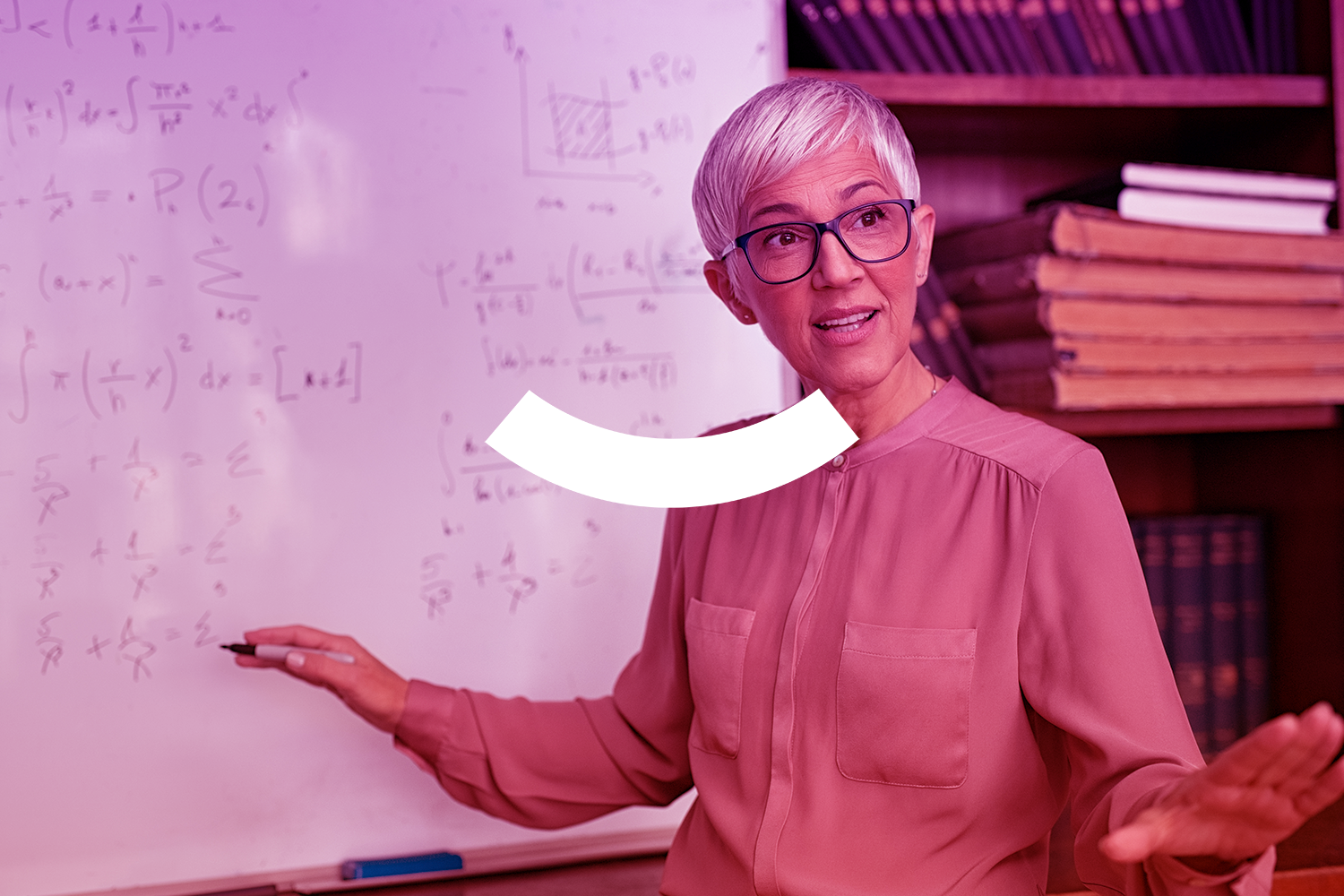 Teacher beside whiteboard, gesturing in a classroom with bookshelves behind her.