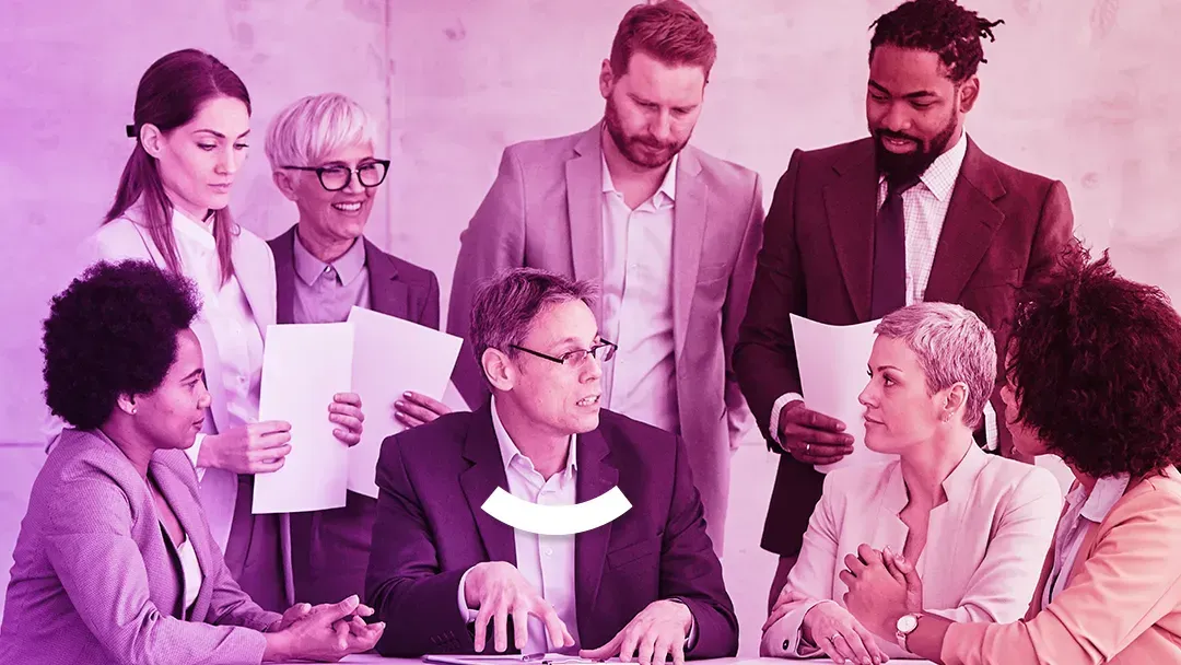 Office team gathered around a laptop, discussing a document in a pink-tinted meeting room.