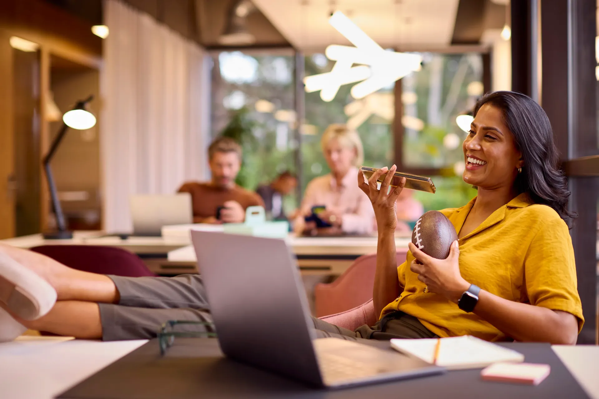 Woman in yellow smiling with her feet up on a table in front of a laptop.