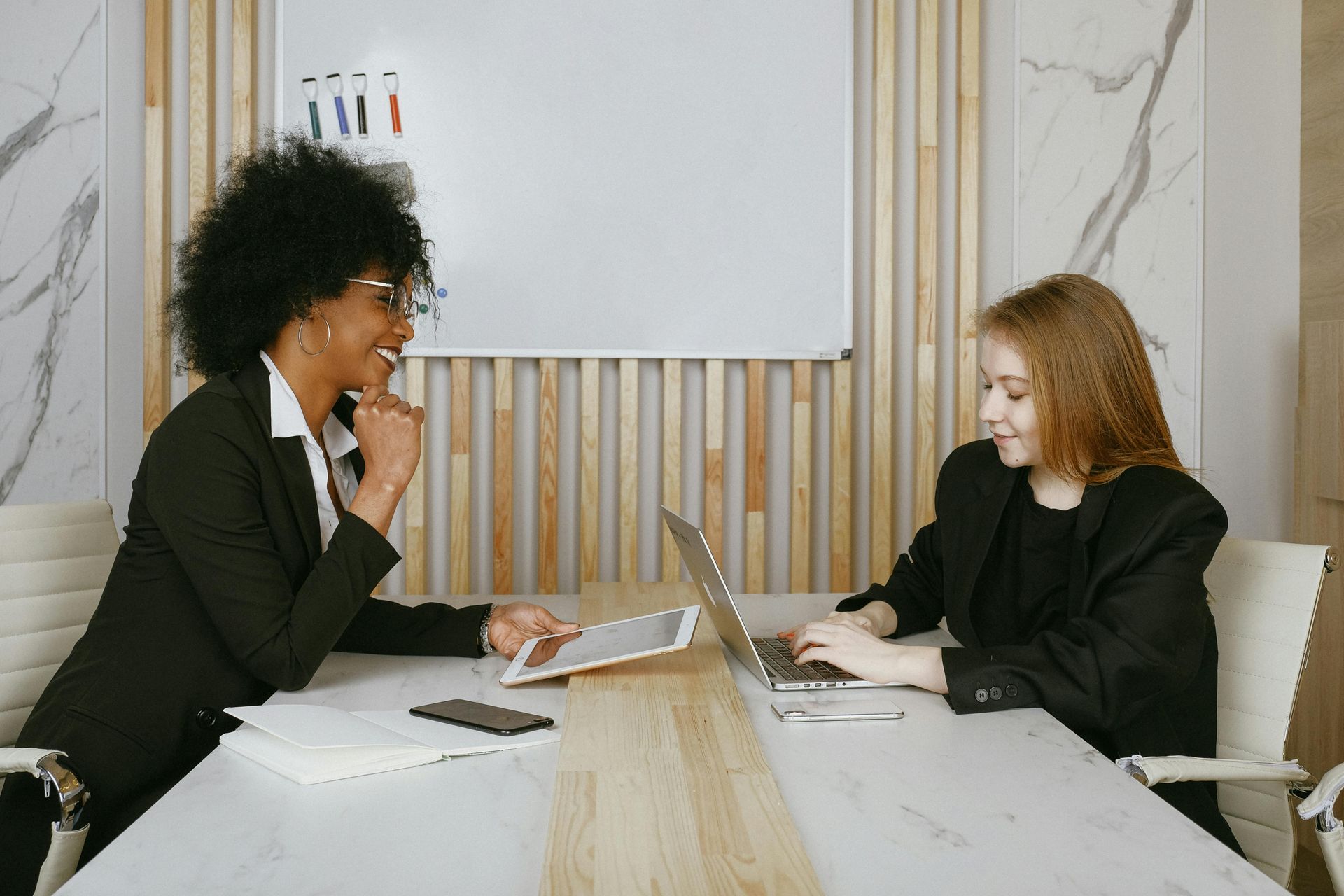 Two people in suits, one with a tablet, the other a laptop, at a table, smiling in an office.