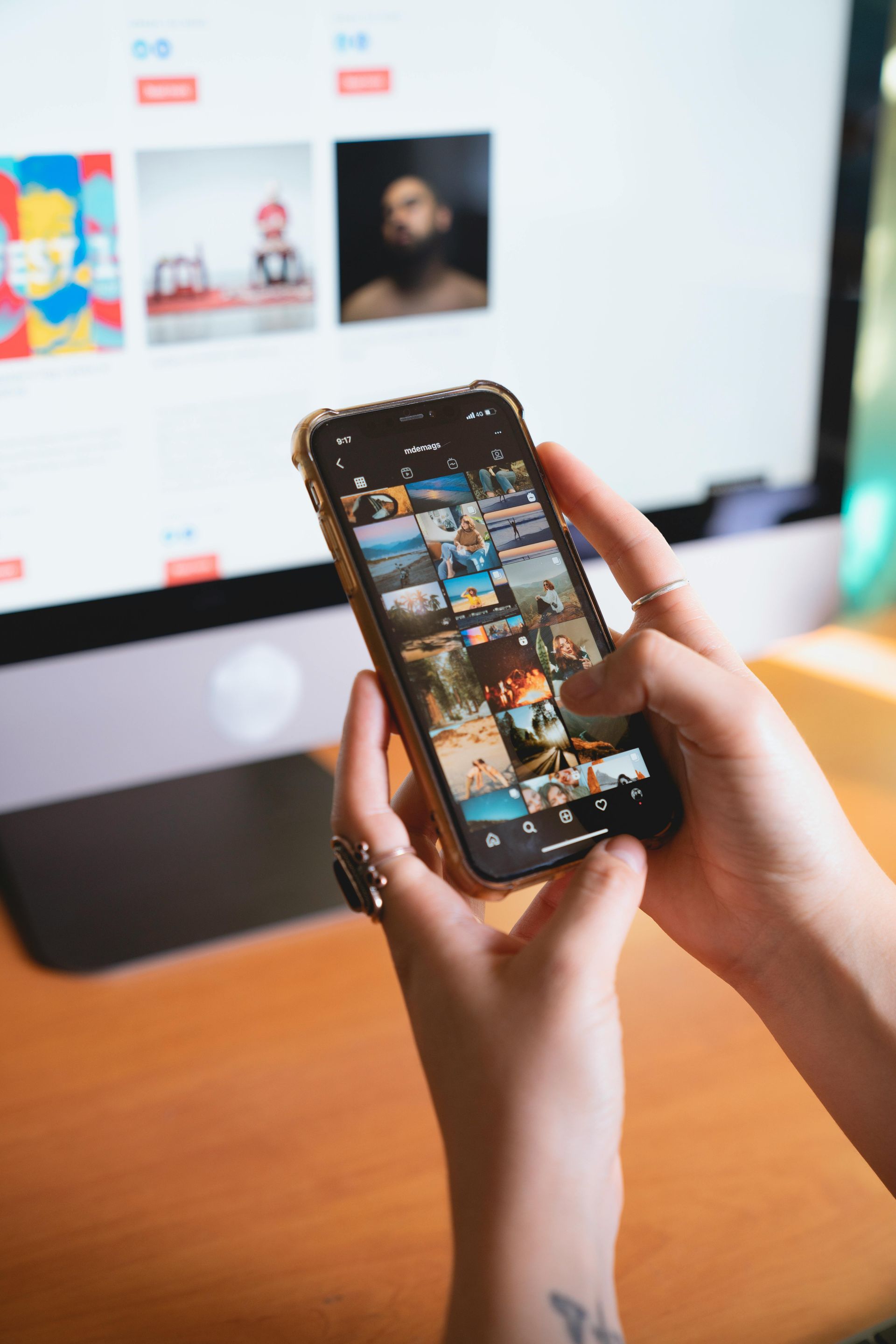 Hands holding a smartphone in front of a desktop monitor, browsing a photo gallery.