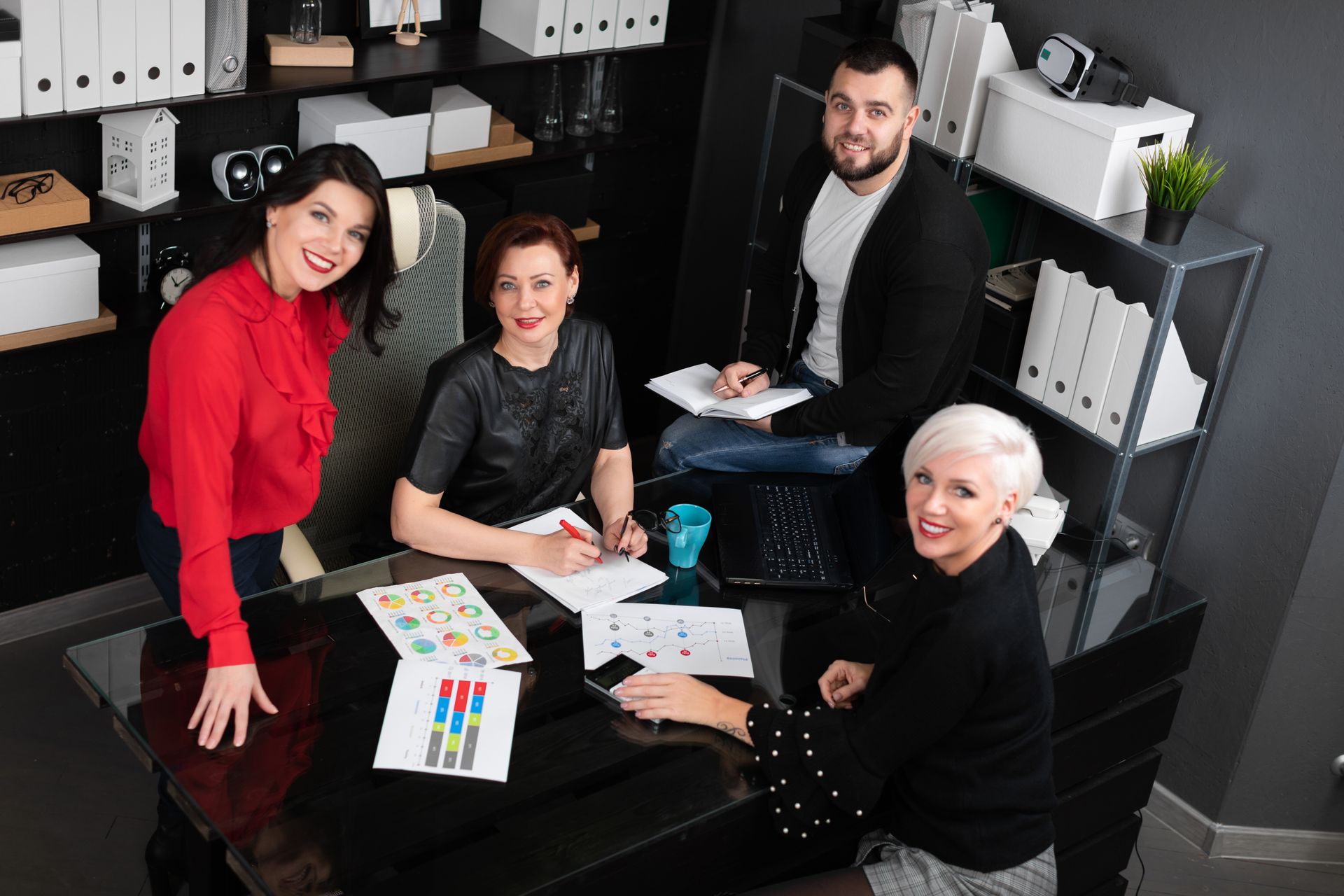 Four people collaborating around a desk in an office, smiling and reviewing documents and colorful charts.
