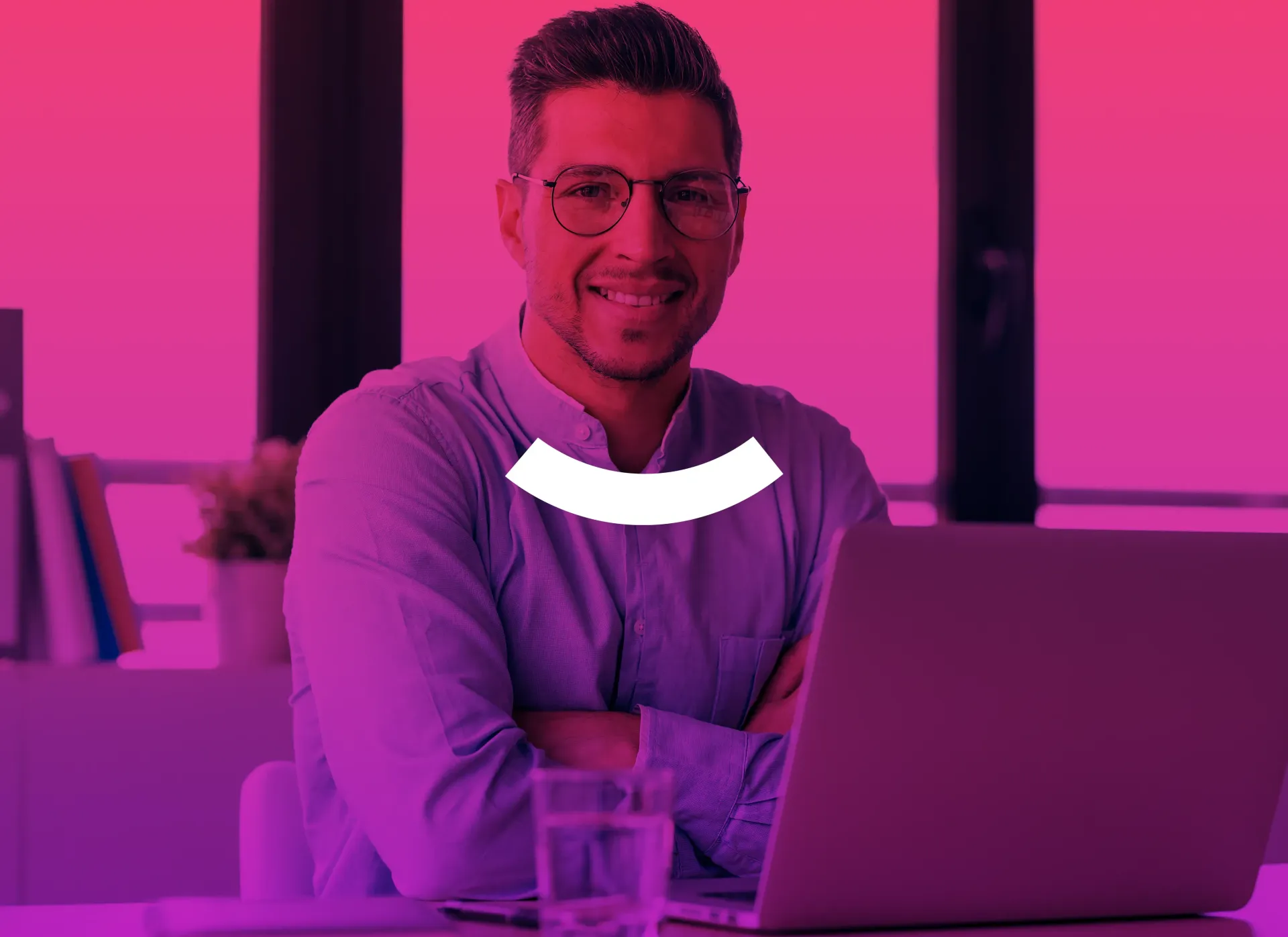 Smiling person at a laptop in a pink-lit office, wearing glasses and a hoodie