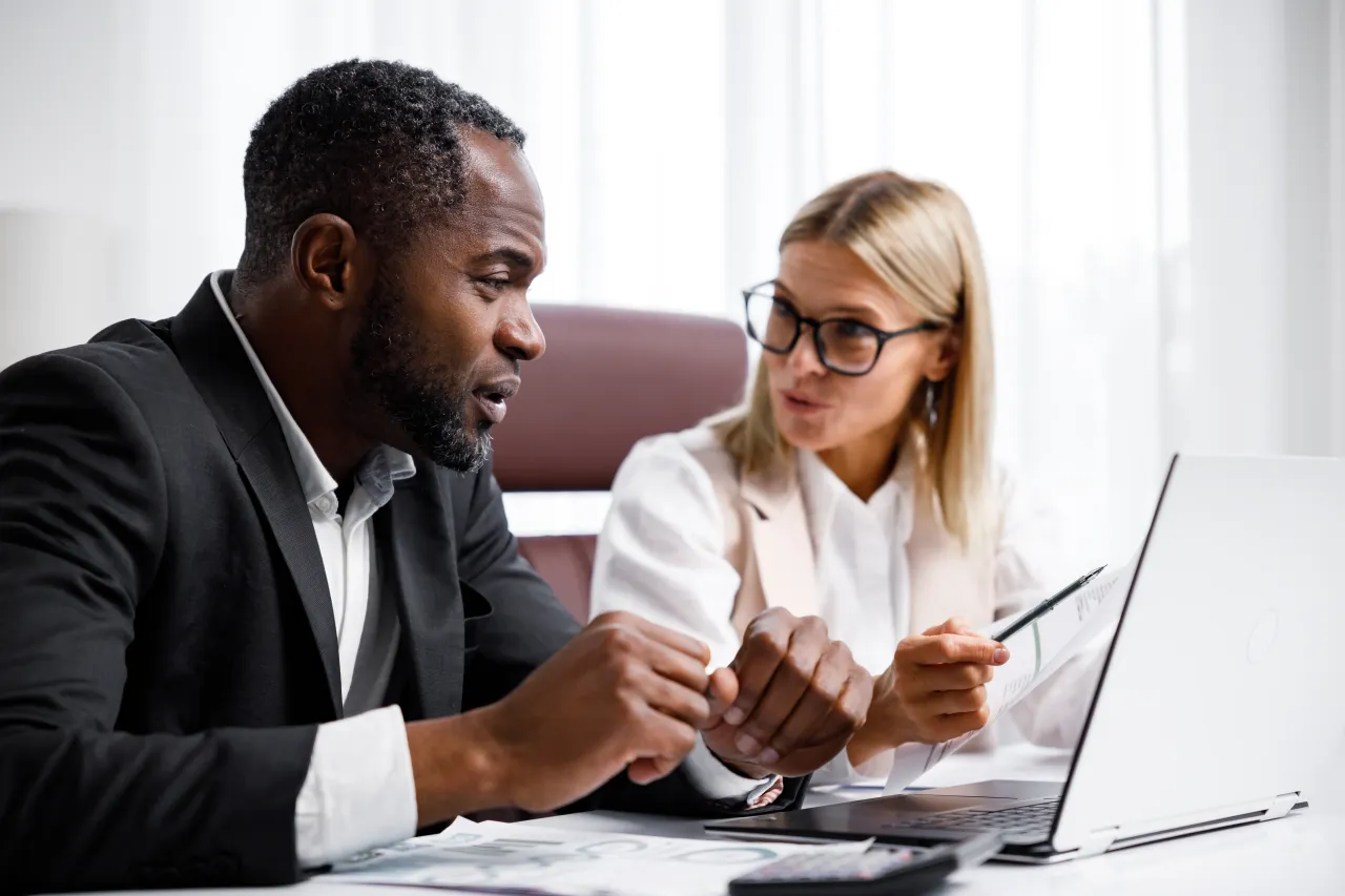 A man and a woman are sitting at a table looking at a laptop.