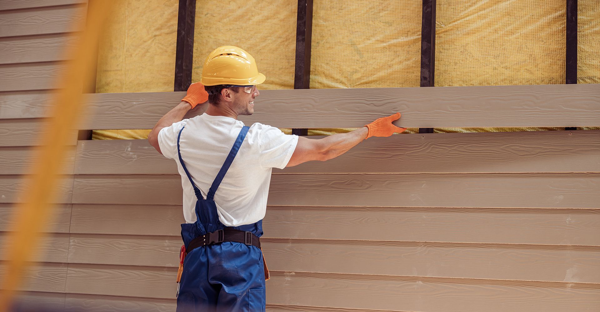 A man wearing a hard hat and overalls is working on a wooden wall.