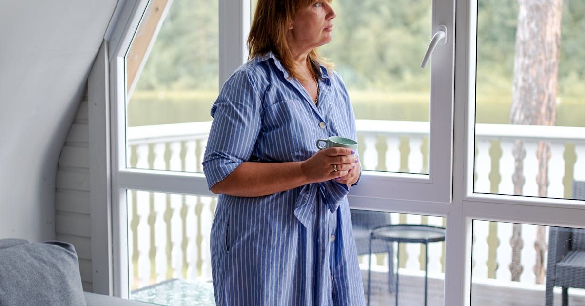 Vrouw in een blauwe jurk met een drankje in haar hand bij een licht raam met uitzicht op een balkon
