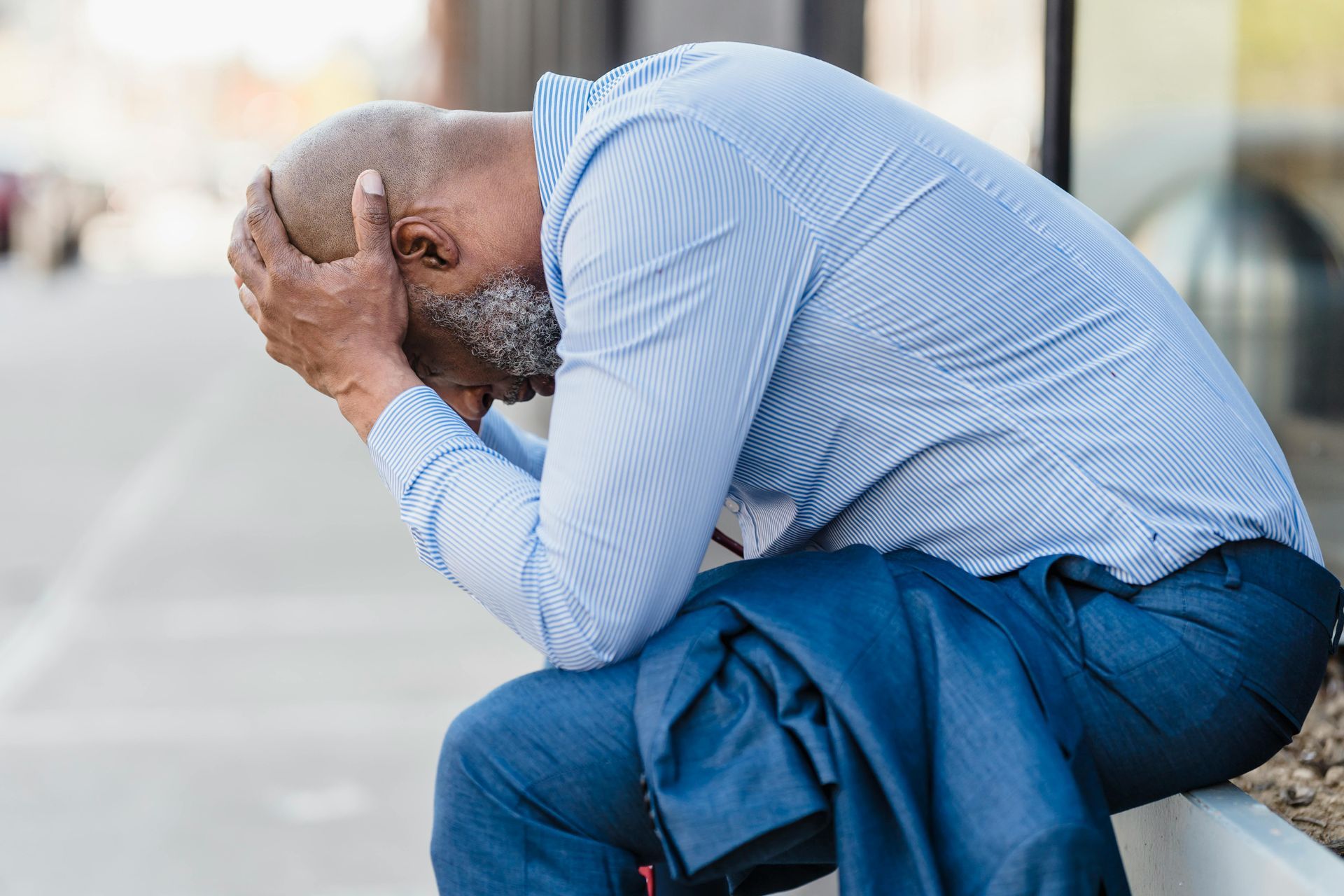 Een man in een blauw shirt zit voorovergebogen met zijn hoofd in zijn handen op een bankje en ziet er bedroefd uit.