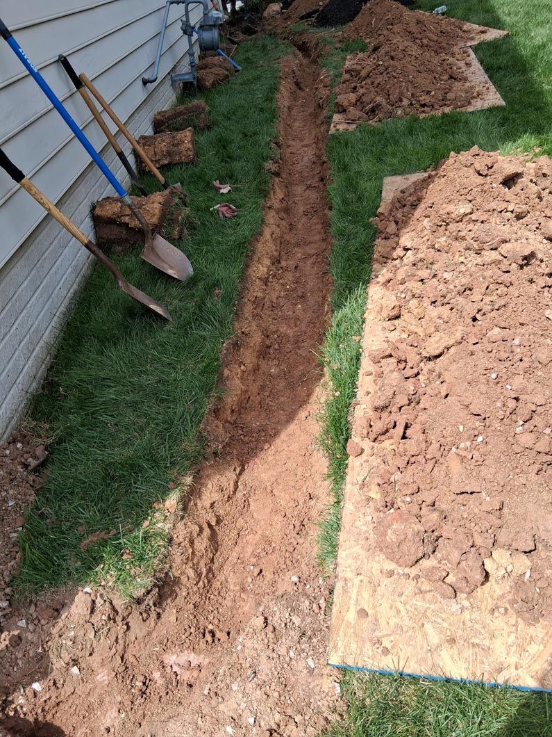 Narrow trench dug beside a house, with shovels and piles of excavated soil along the grass.