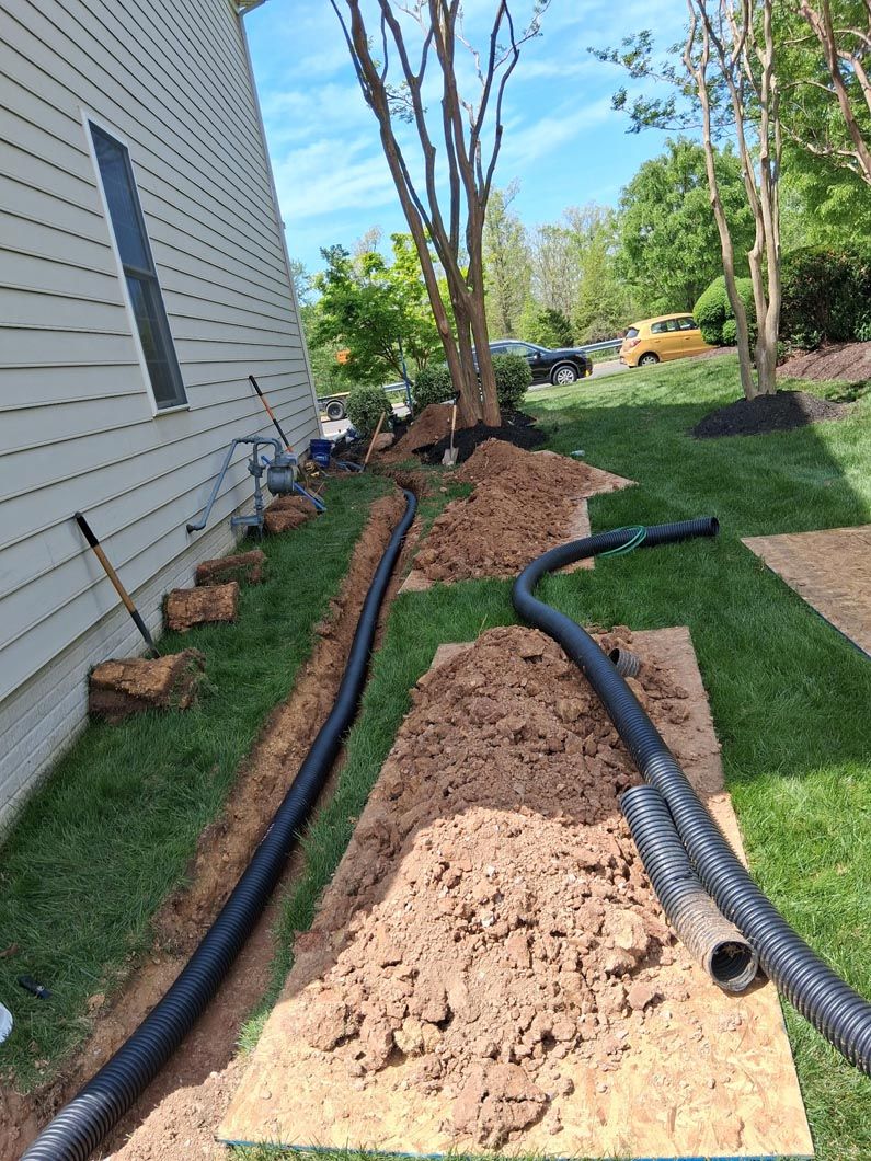 Backyard trench with black drainage pipes and mounds of dirt beside a house under construction