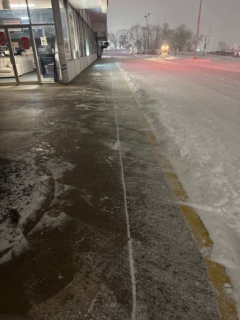 Snowy sidewalk beside a glass-fronted building at night, with slushy road and red taillights in the distance