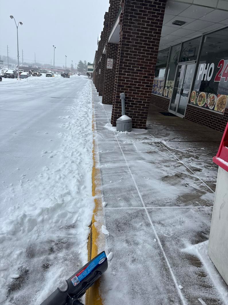 Snowy sidewalk beside a storefront, with snow piled along the curb and a partially cleared walkway.