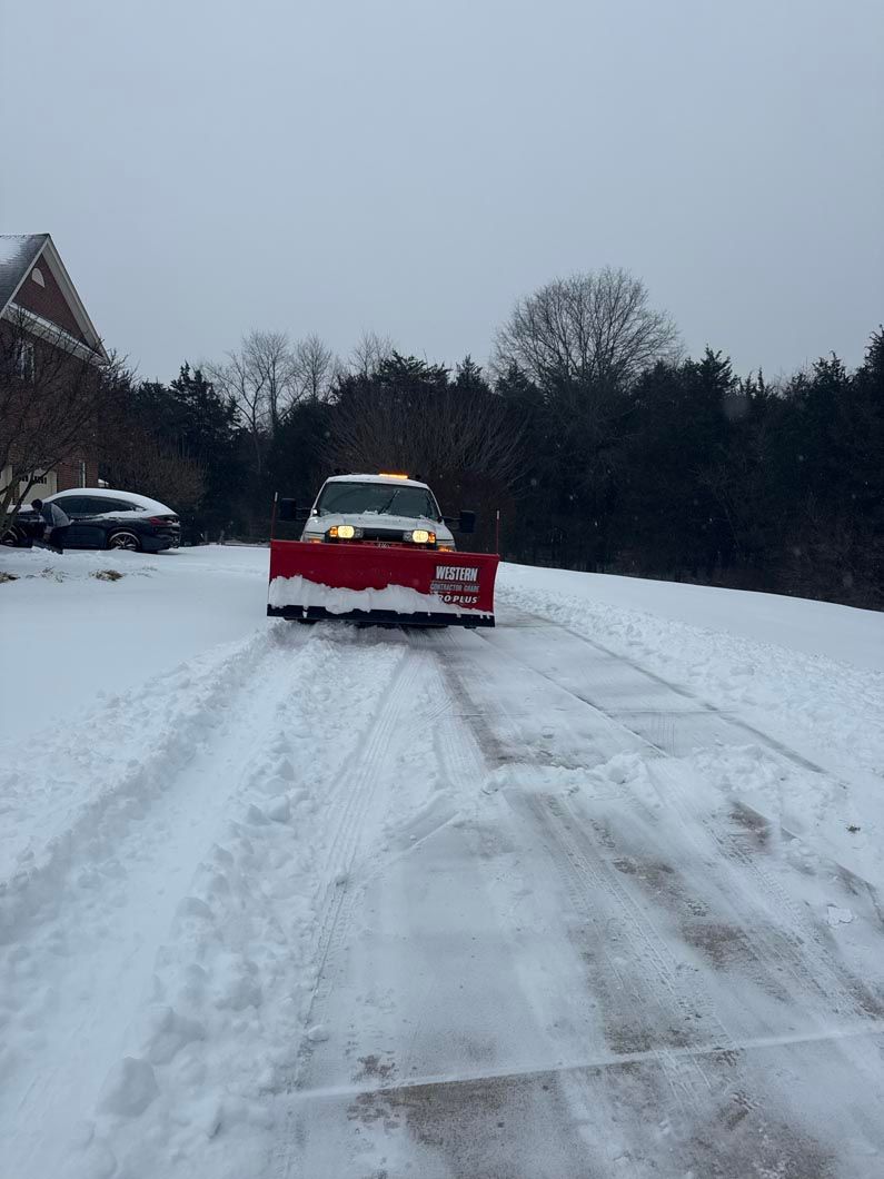 Snow-covered road with a red snowplow truck clearing a driveway at dusk.
