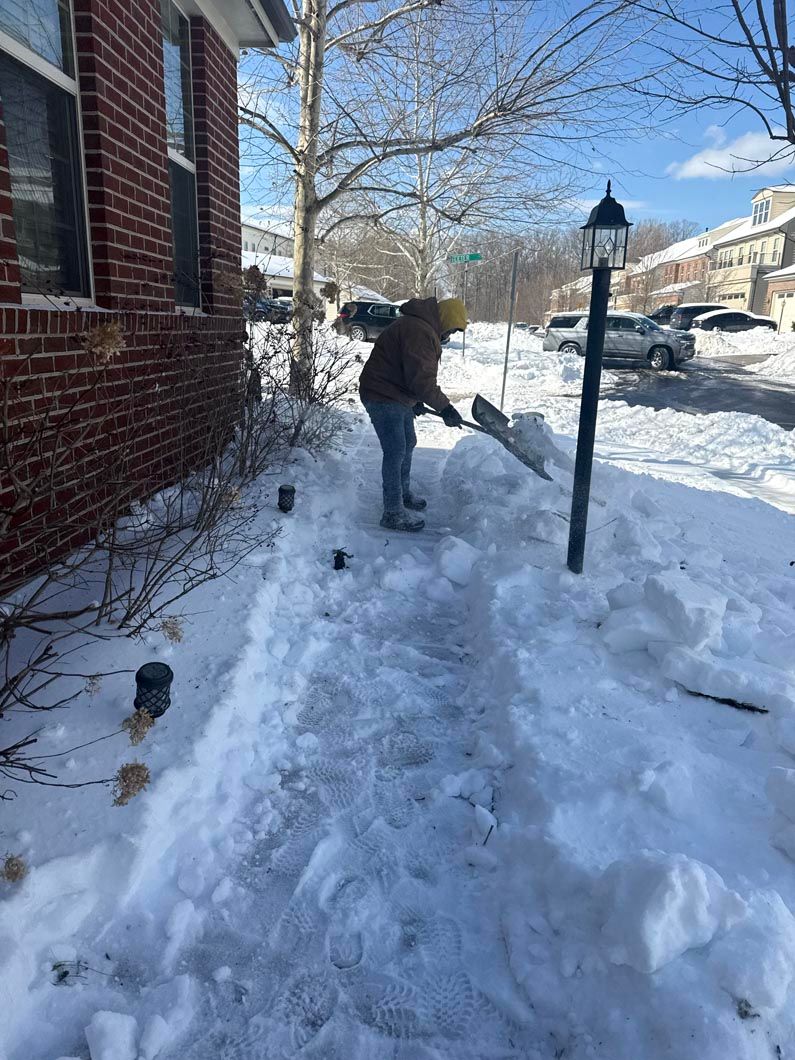 Person shoveling a snow-covered walkway beside a brick house on a sunny winter street