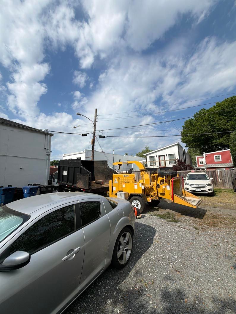 Silver car beside yellow construction equipment in a gravel lot under a cloudy blue sky