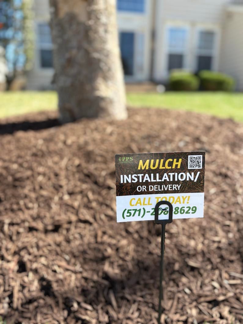Small yard sign for mulch installation/old mulch removal in a landscaped area with blurred building behind.