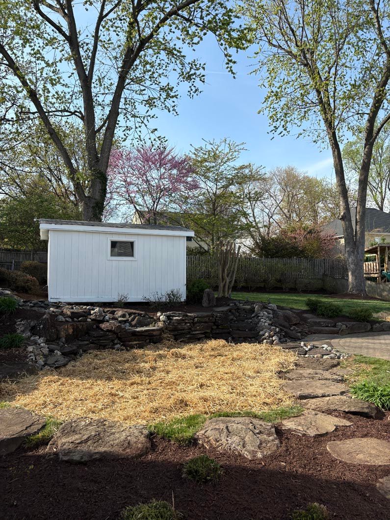 White shed in a wooded yard with a straw-covered patch of ground and trees in the background