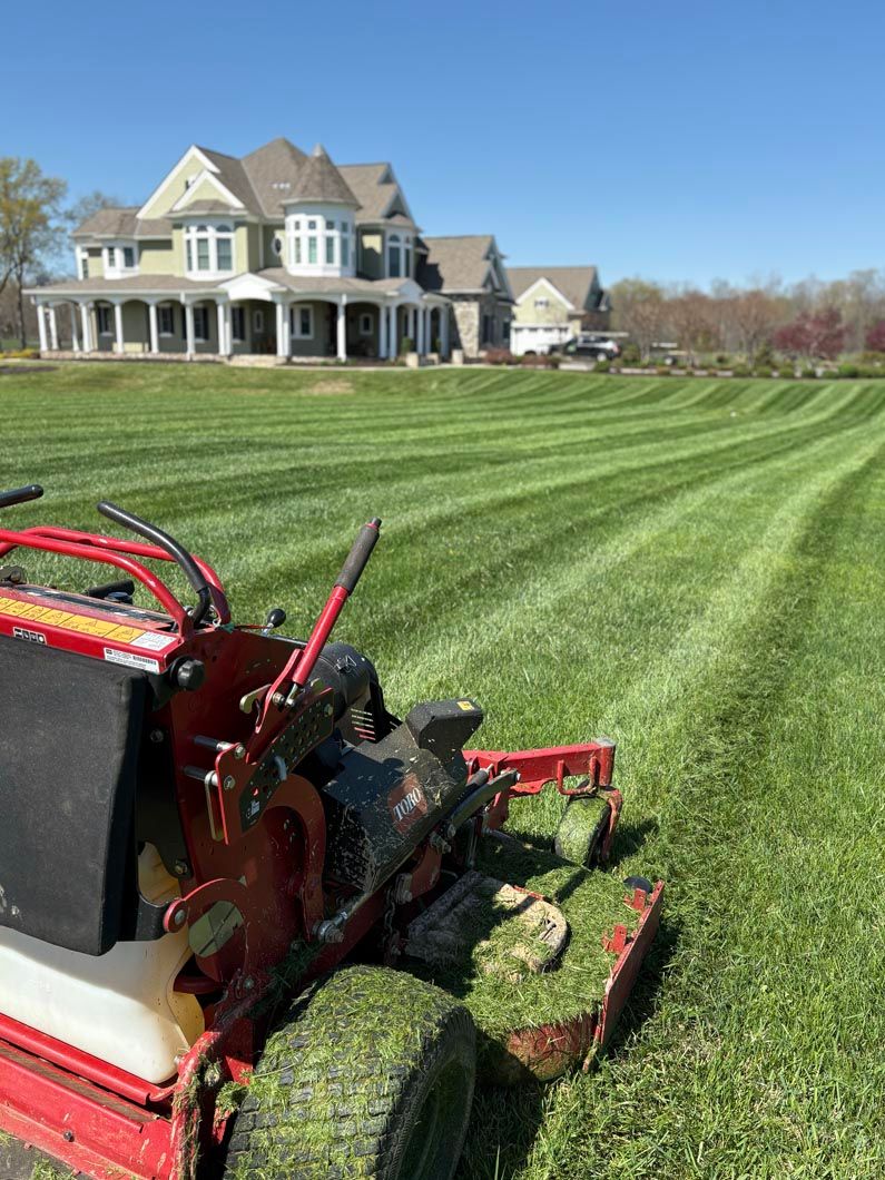 Red lawn mower cutting striped grass in front of a large house on a sunny day
