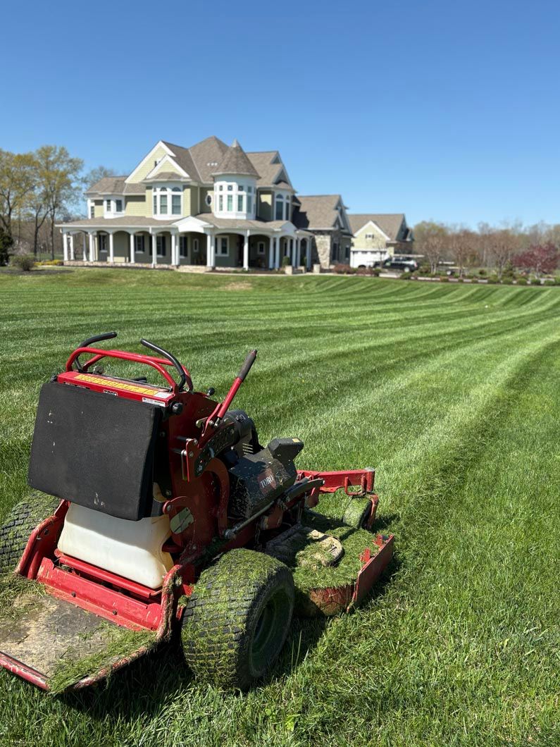Red lawn mower on a striped green field with a large house in the background.