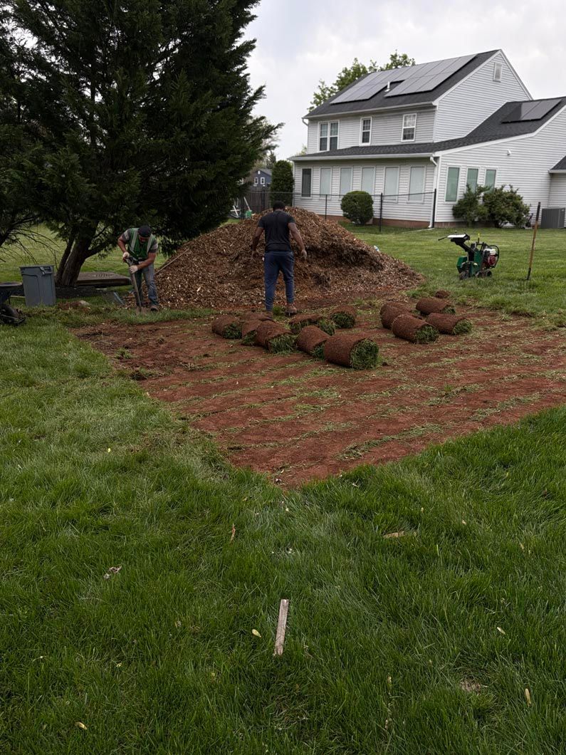 Person standing on mulch-covered garden bed beside a white house and lawn.